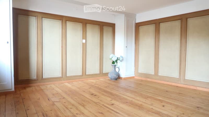 Empty room with wooden flooring, white walls, and large wooden paneling. A single vase with white flowers is placed in the center.
