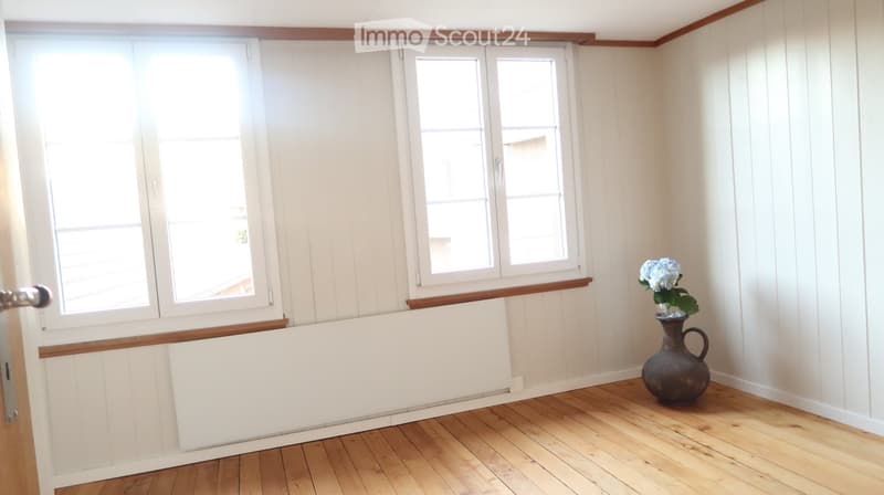 Bright room with wooden flooring, white paneled walls, large windows, and a decorative vase with flowers on the floor.