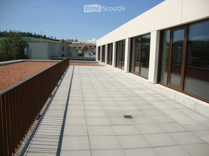Modern rooftop terrace with light grey tiled flooring, metal railing, and large windows overlooking a residential area.