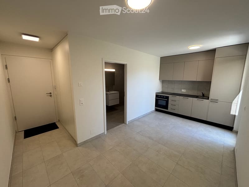 Empty hallway with tiled floor, white walls, and a small kitchen area visible through an open doorway. The kitchen features grey cabinets, a sink, and an oven.