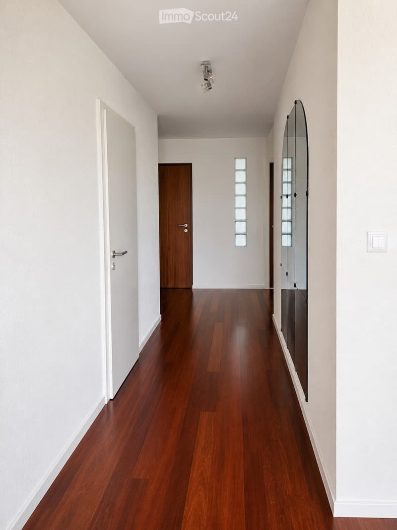 Clean, modern hallway with polished wooden flooring, white walls, and recessed lighting. Features a large mirror on one wall and several doors leading to other rooms.
