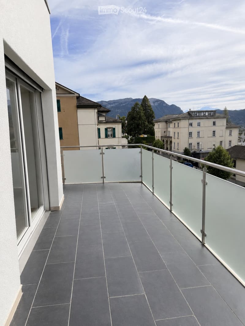 Modern balcony with grey tiled flooring, glass railing, and a view of surrounding buildings and mountains.