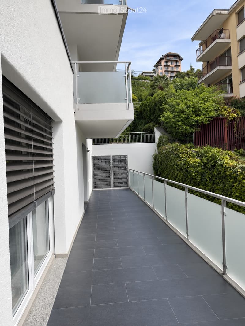 Modern balcony with dark grey tiled flooring, glass railing, and metal balustrade. The balcony is adjacent to a white building with large windows and shutters. The background shows greenery and neighboring buildings.