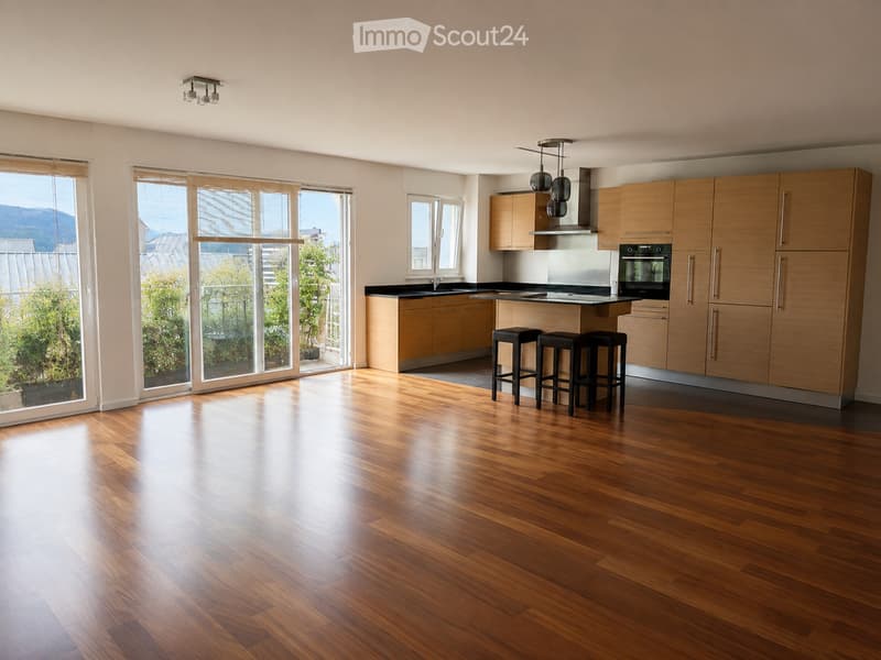 Modern kitchen with light wood cabinets, stainless steel appliances, island with bar stools, and large windows overlooking a garden.