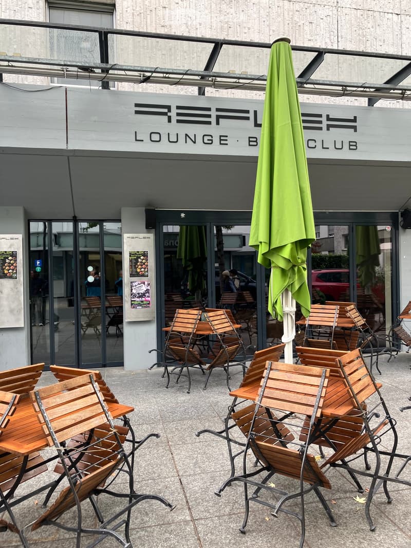 Outdoor seating area with wooden chairs and tables, a green umbrella, and a sign for "Reflex Lounge Club" above the entrance.