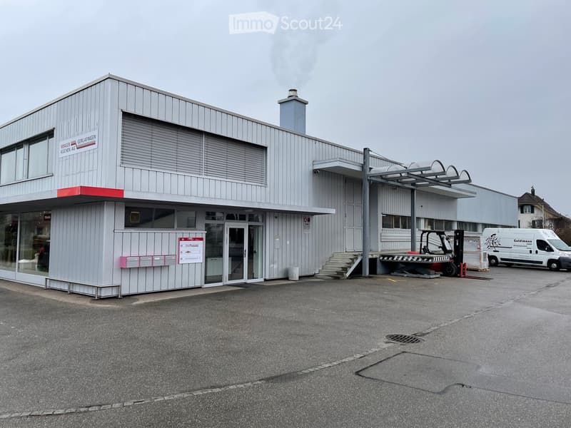 Modern industrial-style building with a grey metal facade, large windows, and a small chimney. Features include a covered entrance area, a small staircase leading to a raised platform, and a parking area with a forklift and van.