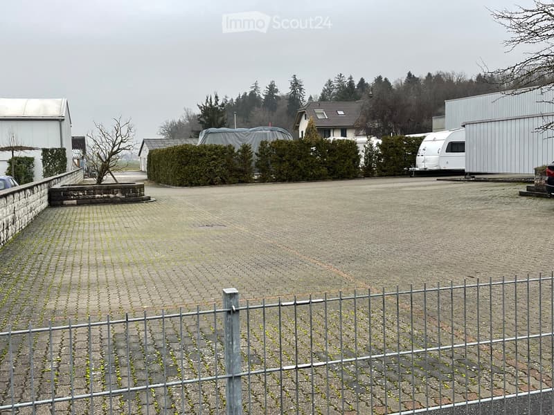 A paved outdoor area enclosed by a metal fence, with a small garden bed, a covered vehicle, and surrounding buildings.
