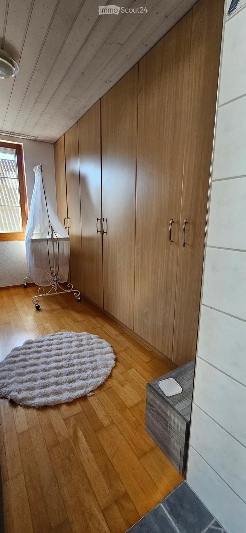 Spacious attic room with wooden flooring, large built-in wardrobes, a round shaggy rug, a small stool, and a baby crib with a canopy.