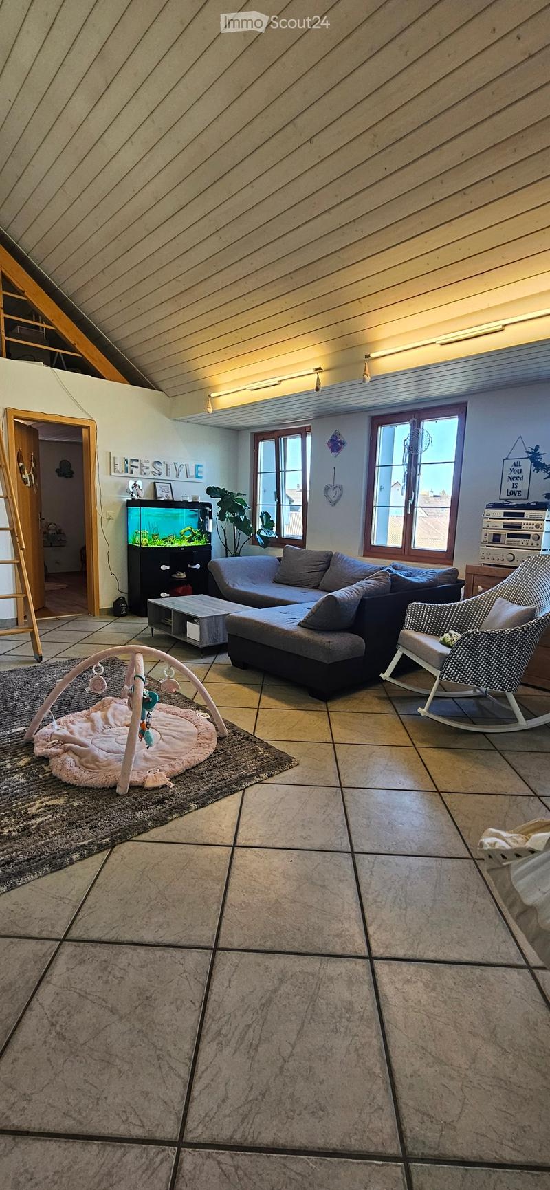 Bright living room with a grey sectional sofa, coffee table, rocking chair, fish tank, baby playpen, and a plant near the windows.