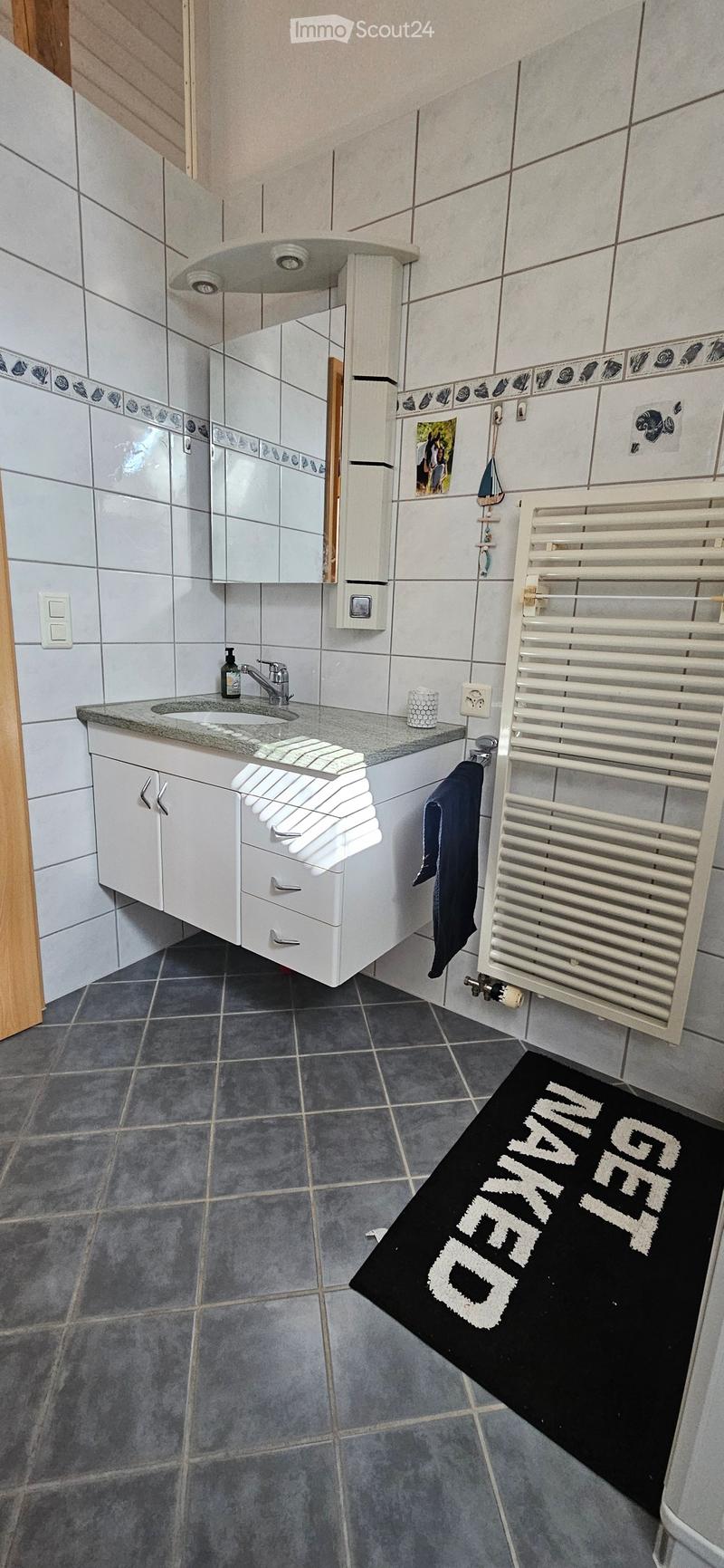 Modern bathroom with white tiled walls, grey tiled floor, white sink cabinet, mirror, radiator, and a black bathroom rug with white text.