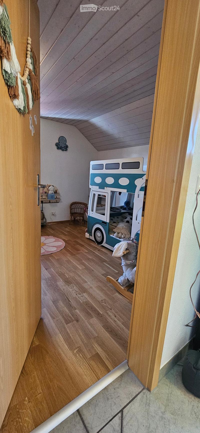 Cozy children's bedroom with wooden floors, slanted ceiling, toy van bed, wicker chair, rocking horse, and a small shelf with toys.