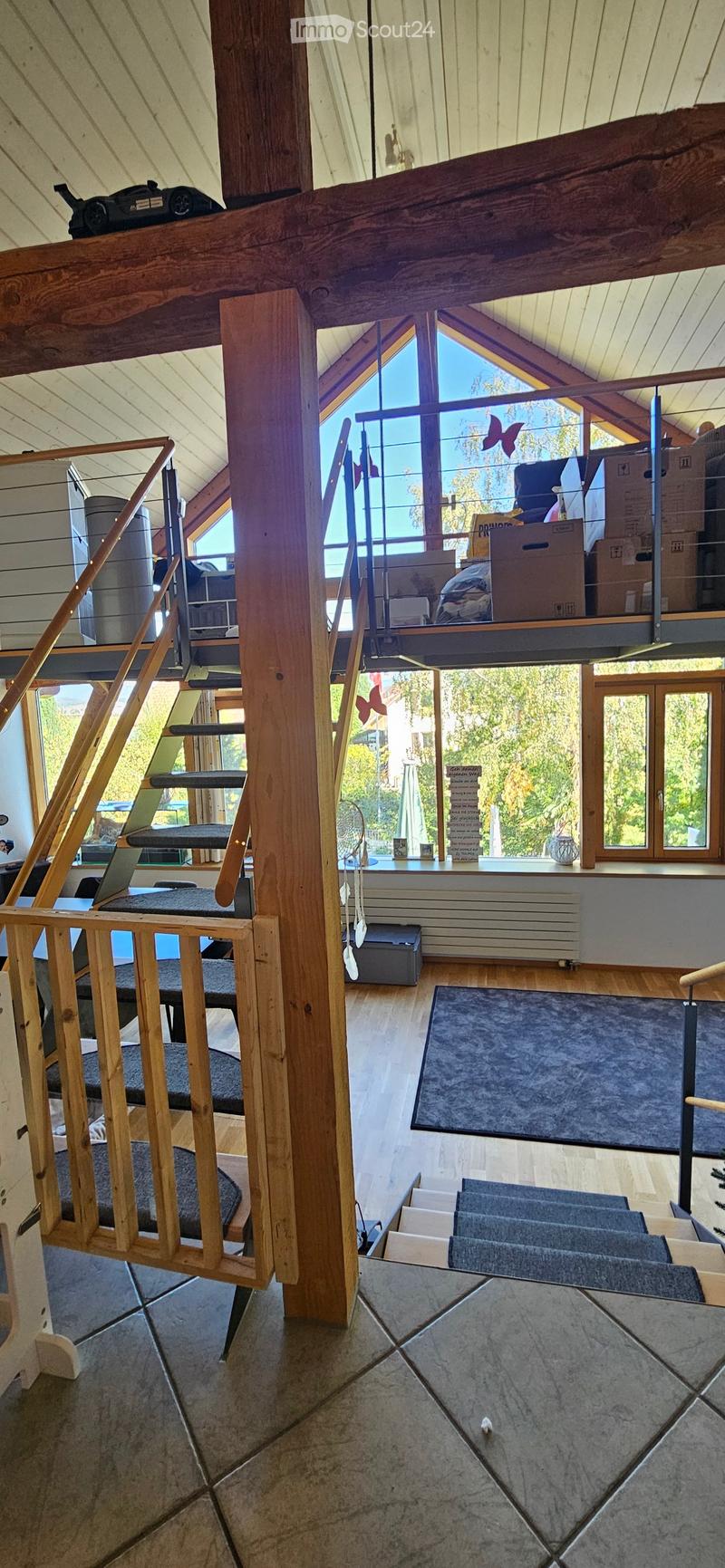 The image shows a modern, open-concept living space with wooden beams and large windows. Visible objects include a staircase, a cat tree, a laptop, a fan, a radiator, a table, a chair, and a toy car. The room features a mix of wooden and tiled flooring, w