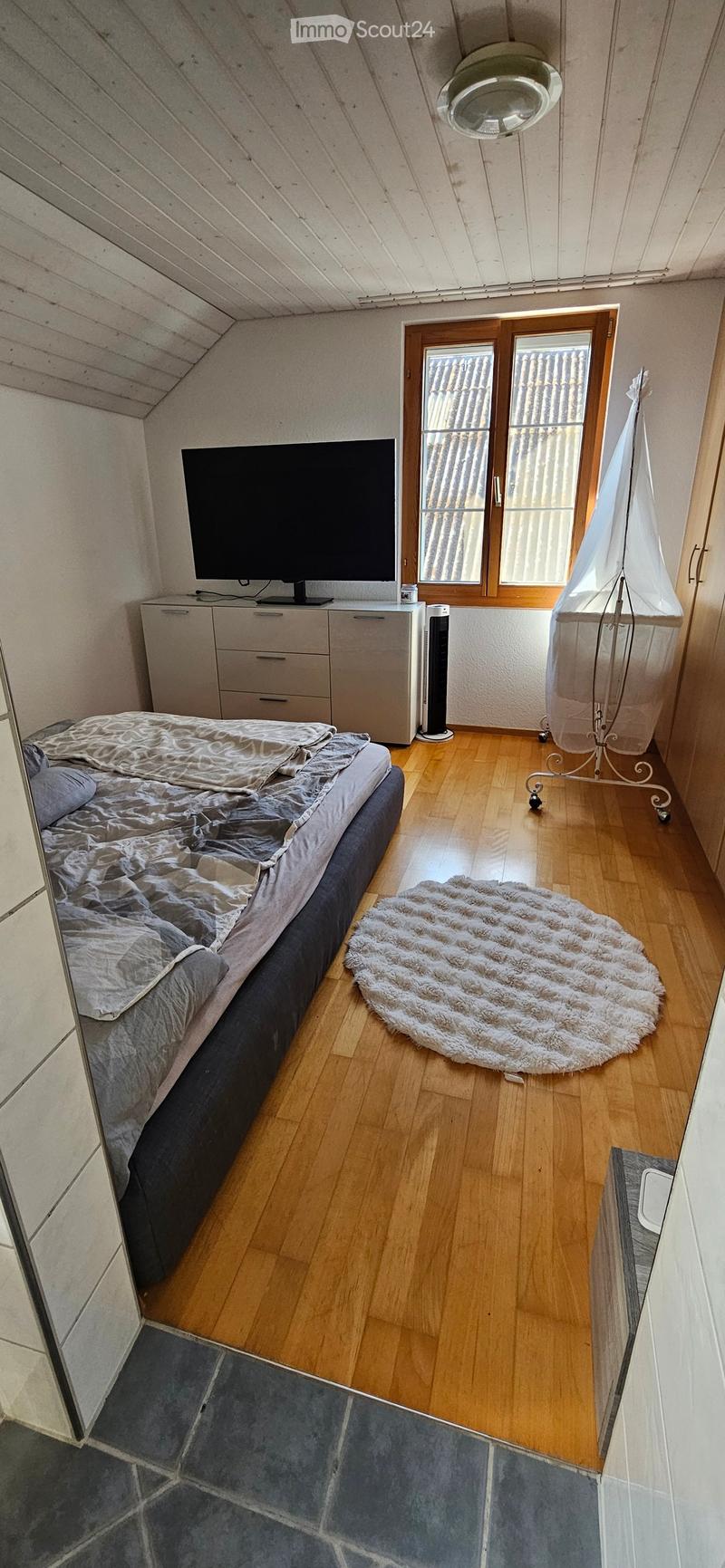 Cozy attic bedroom with wooden floors, slanted ceiling, bed, television, cabinet, fan, and a round fur rug.
