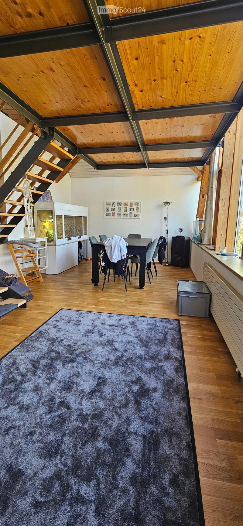 A spacious, open-concept living and dining area with wooden floors, a large grey shag rug, a dining table with chairs, a speaker, and a staircase leading to an upper level.