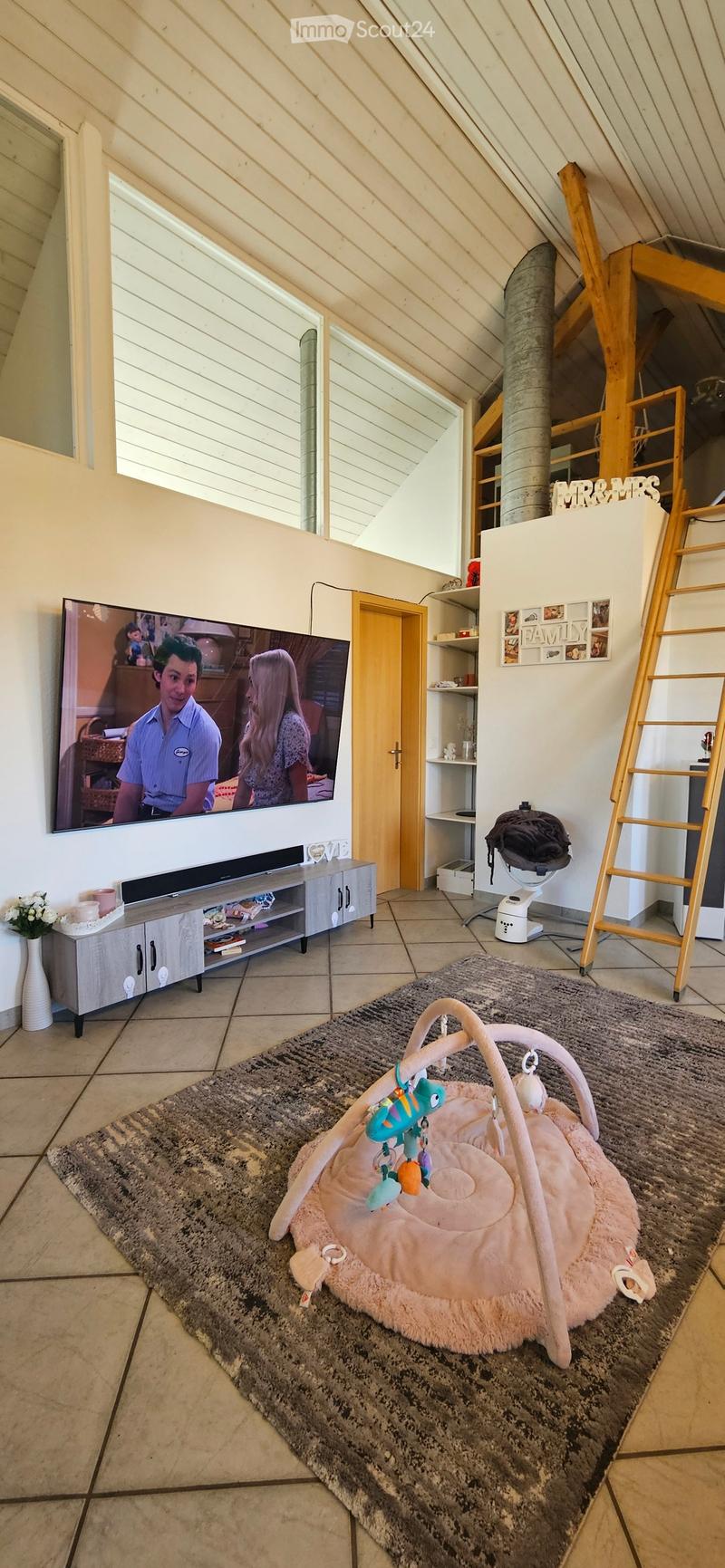 A cozy living room with a television, cabinet, baby seat, vase, shelves, and a ladder. The room features a slanted ceiling and large windows.