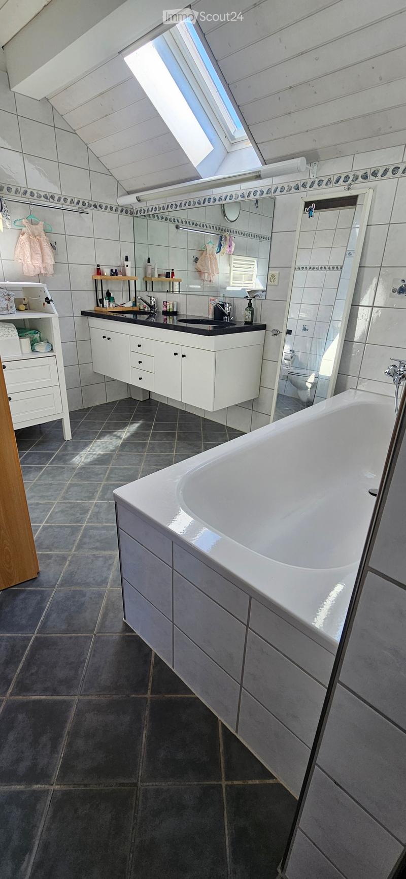 Modern bathroom with a large bathtub, double sink, mirror, and built-in cabinets. The room features dark tiled floors and walls with a skylight window providing natural light.