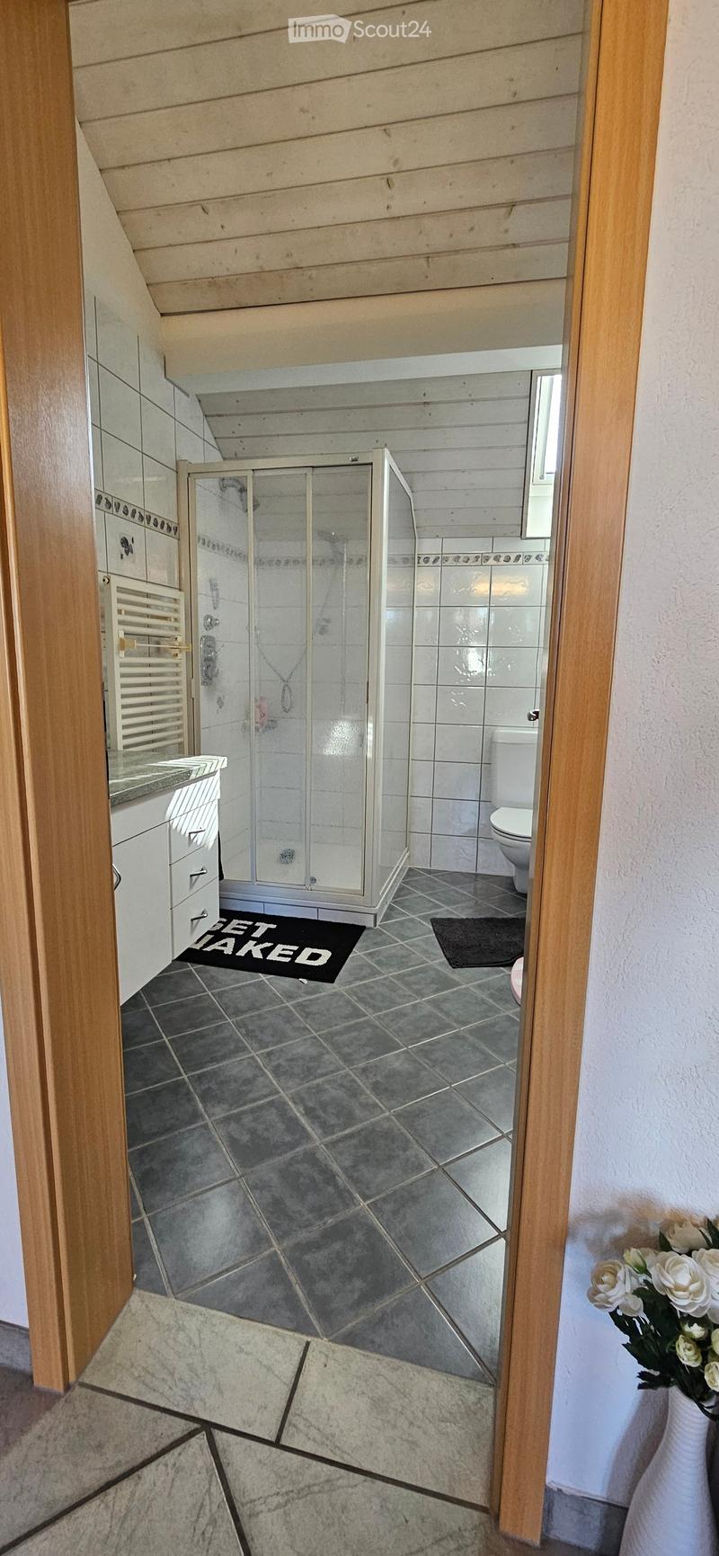 Modern bathroom with a glass-enclosed shower, toilet, sink, and radiator. The floor is tiled, and there is a black bathroom mat in front of the shower.