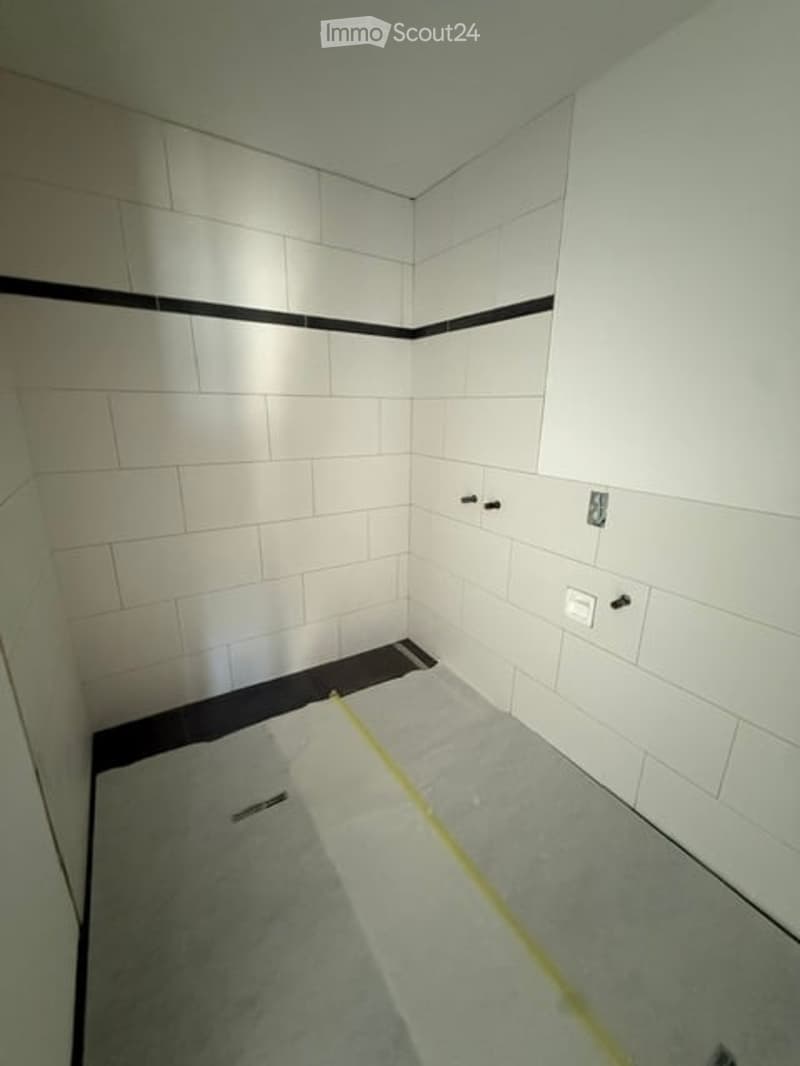 Empty bathroom with white tiled walls and floor, featuring a black border along the upper wall and a gray floor. The space appears to be under renovation or construction, with no fixtures or furniture present.