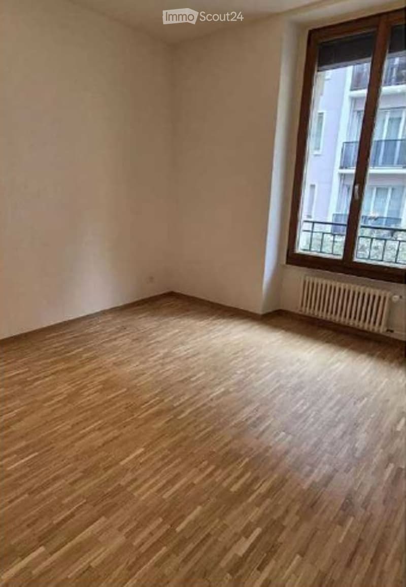 Empty room with wooden flooring, white walls, and a large window with a radiator below it.