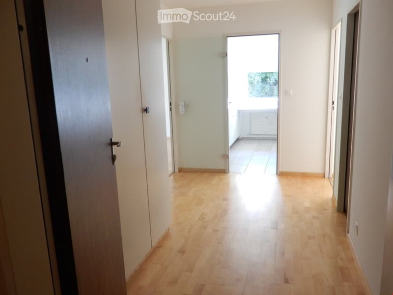 Empty hallway with light wooden flooring, white walls, and a glass door leading to a balcony area.