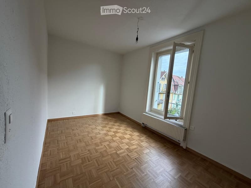 Empty room with wooden herringbone flooring, white walls, and a large window with a radiator below it. The room has a single light fixture hanging from the ceiling.