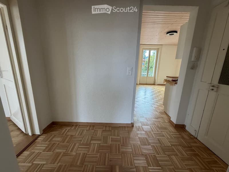 Empty hallway with wooden flooring and white walls, leading to an open area with a visible kitchen and balcony.