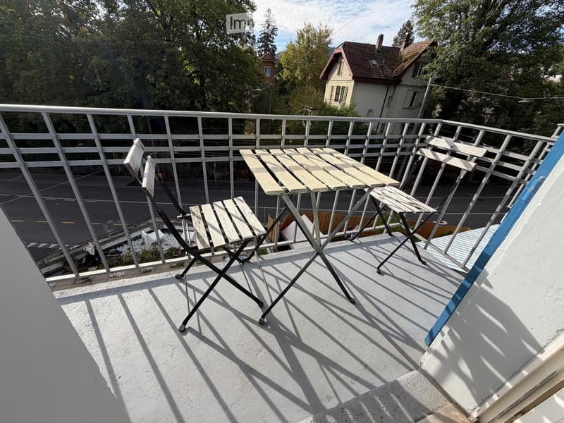 Balcony with a small wooden table and two folding chairs, overlooking a street with trees and houses.