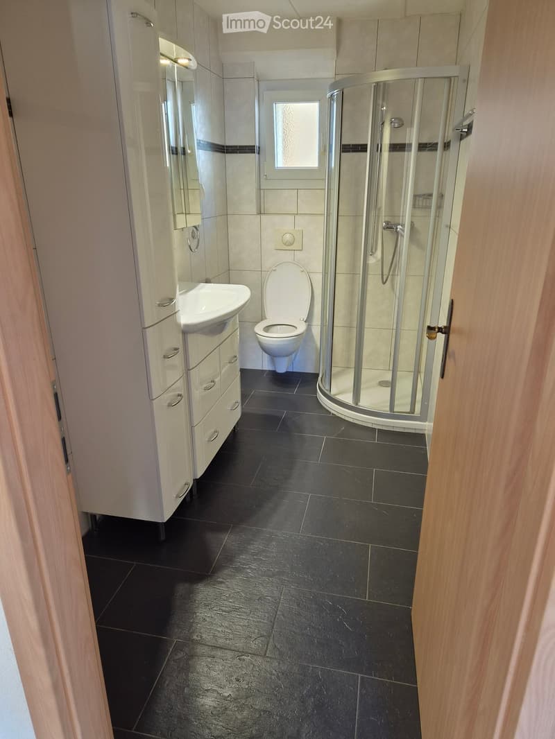 Modern bathroom with black tiled floor, white sink, toilet, and glass-enclosed shower. Features a white cabinet with drawers and a small window above the toilet.