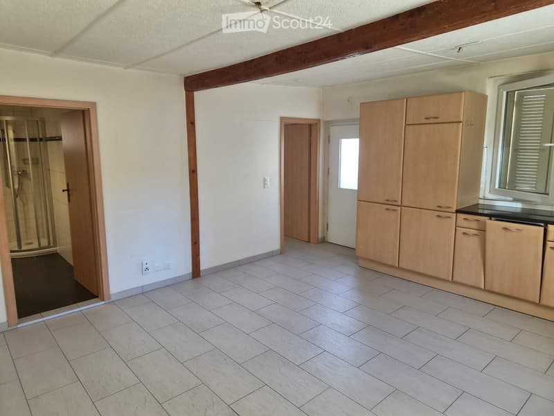 Empty room with tiled floor, white walls, wooden cabinets, and a glass-enclosed shower area visible through an open doorway.