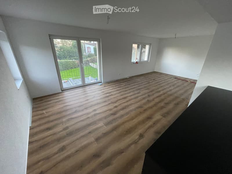 Empty room with wooden flooring, white walls, large sliding glass door leading to a garden, and a window on the opposite wall.