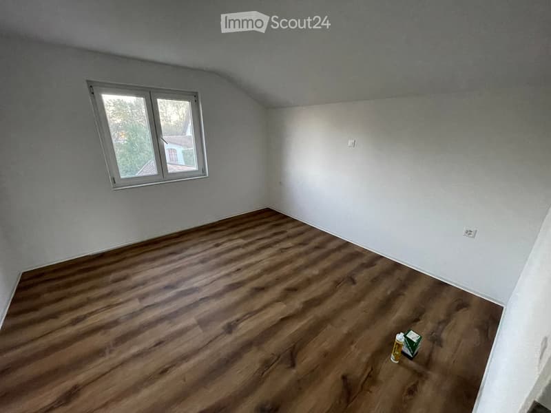 Empty room with wooden flooring and white walls, featuring a single window with white frame and a small electrical outlet visible on the wall.