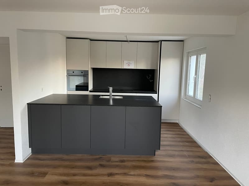 Modern kitchen with grey cabinets, black countertop, island, sink, and oven. Wooden flooring and white walls.