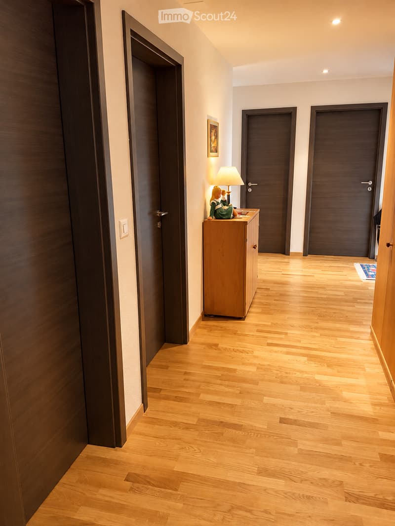 A well-lit hallway with wooden flooring, several dark wooden doors, a small wooden cabinet with a lamp, and a decorative item on top.