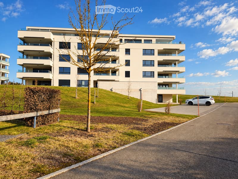 Modern multi-story residential building with balconies and large windows, surrounded by a landscaped area with a tree and a parked car.