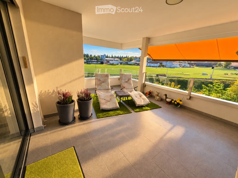 Balcony with two lounge chairs, a small table, potted plants, and decorative items, offering a view of a green field and buildings.