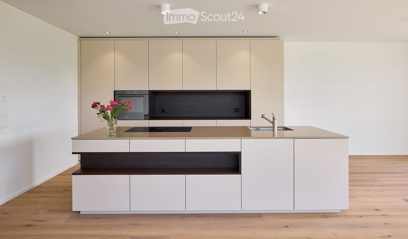 Modern kitchen with white cabinets, a central island, built-in oven, and a sink with a modern faucet. A vase with pink flowers is placed on the island.