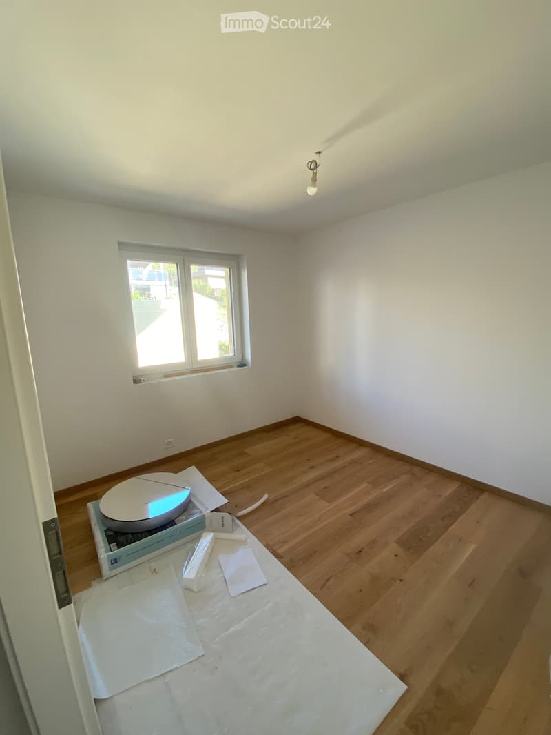 Empty room with wooden flooring and white walls, featuring a small table with a laptop and some papers on it.