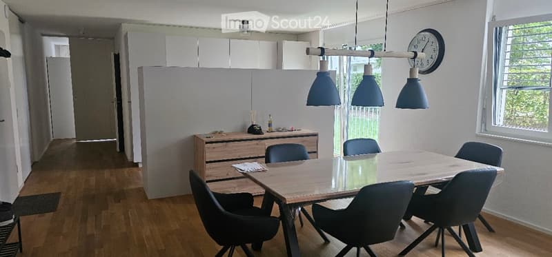 A modern, well-lit room with a wooden dining table surrounded by chairs, a wooden cabinet with various items, and a clock on the wall. The room features wooden flooring and large windows allowing natural light to fill the space.