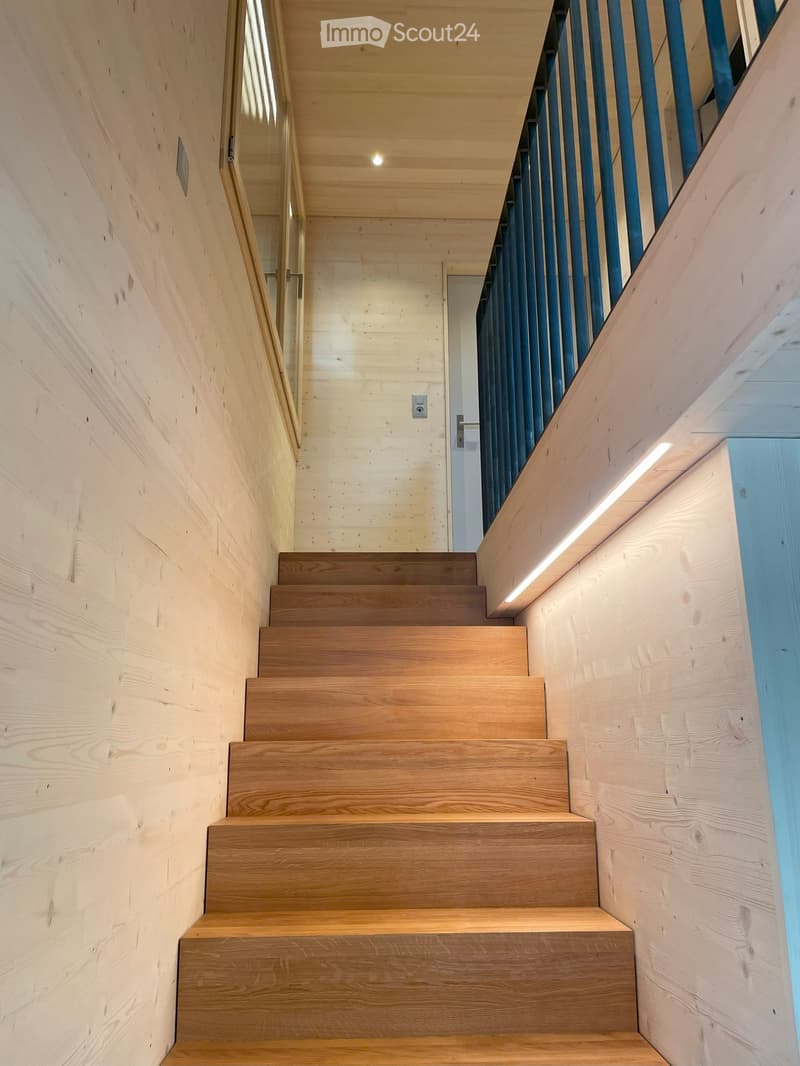 Modern wooden staircase with sleek design, featuring light wood steps and railings, illuminated by integrated LED lighting on the wall.