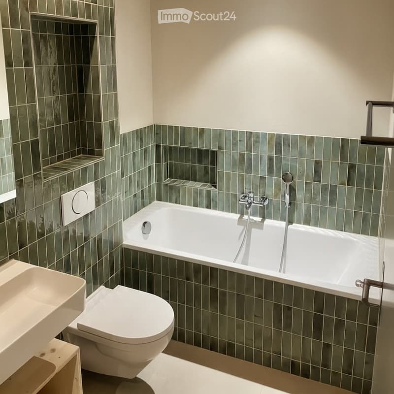 Modern bathroom with green tiled walls, a white bathtub, toilet, and sink. Features a sleek faucet and built-in shelves.