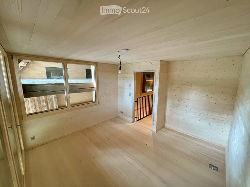 Empty room with wooden flooring, exposed wooden ceiling, and large window with a view of a wooden deck. There is a doorway leading to another room with a staircase.