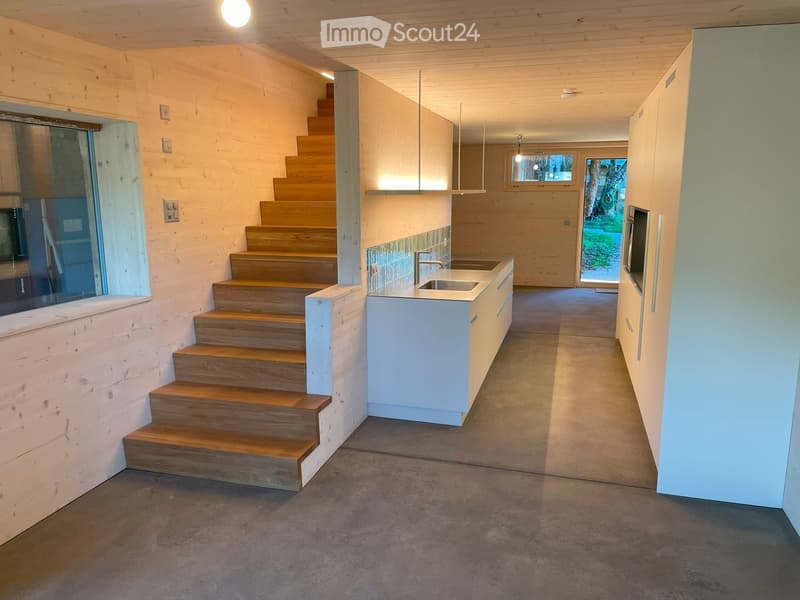 Modern hallway with wooden staircase, white cabinets, and a sink area. Exposed wooden beams on the ceiling and large windows provide natural light.