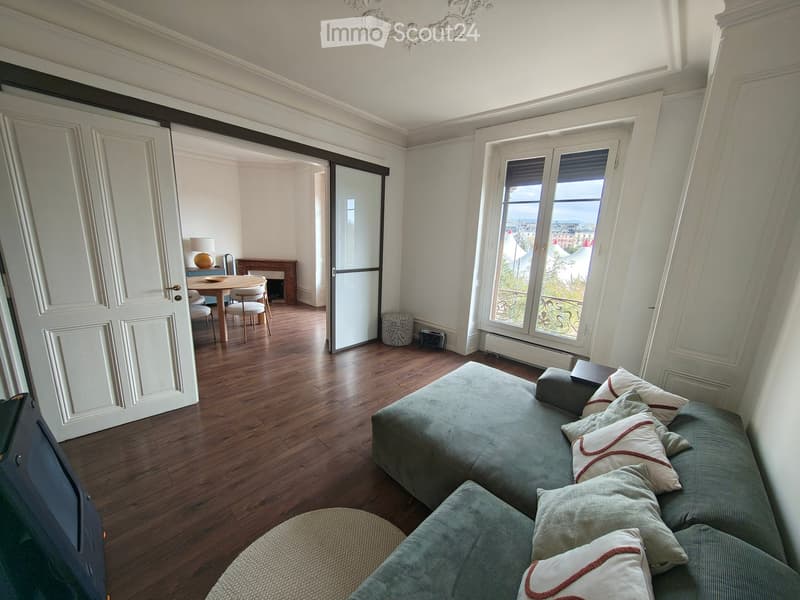 Elegant living room with a grey sectional sofa, decorative pillows, a round wooden coffee table, dining area with chairs, fireplace, and large windows offering a scenic view.