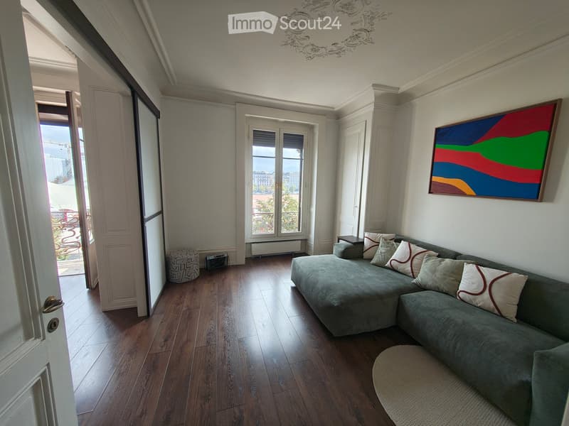 Living room with a grey sectional sofa, ottoman, small table, and colorful painting on the wall. The room has wooden flooring and large windows offering a view.