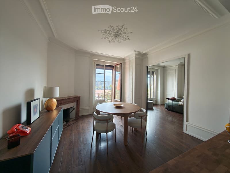 Elegant living and dining area with wooden flooring, a round dining table with chairs, a sideboard with a lamp and picture frame, and a view through large windows.