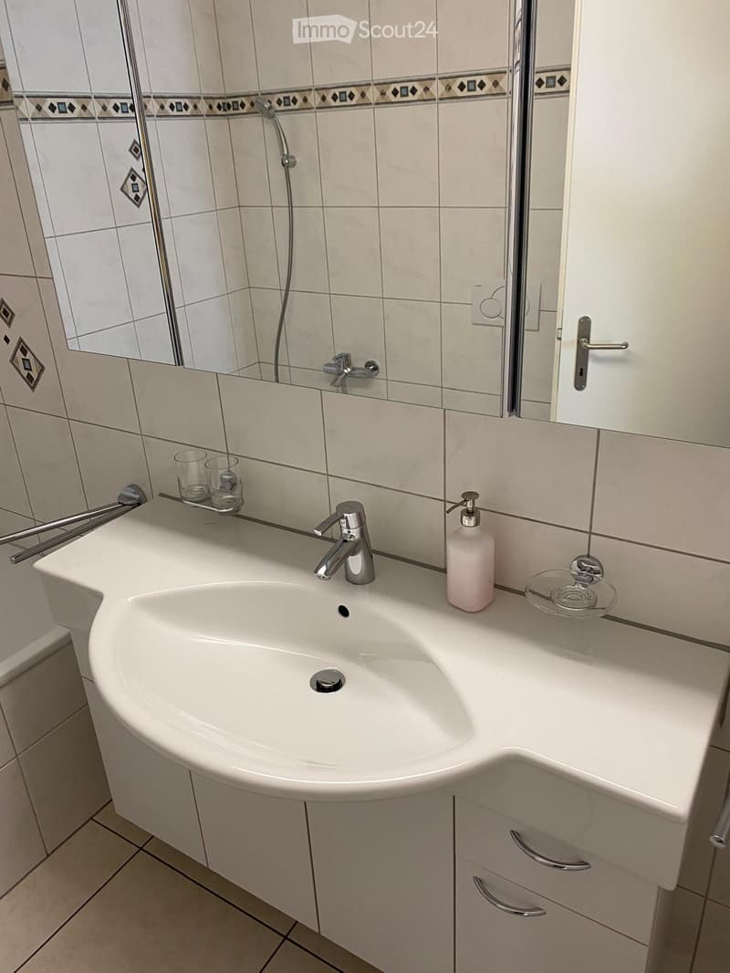 Modern bathroom with a white sink, chrome faucet, soap dispenser, and two cups on the counter. The walls are tiled with a decorative border, and a mirror is mounted above the sink.