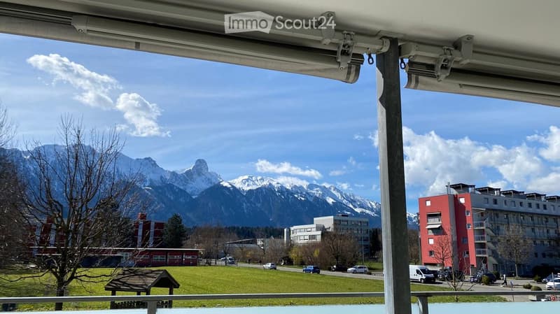 Balcony overlooking a scenic view of mountains, a grassy field, and buildings in the distance.