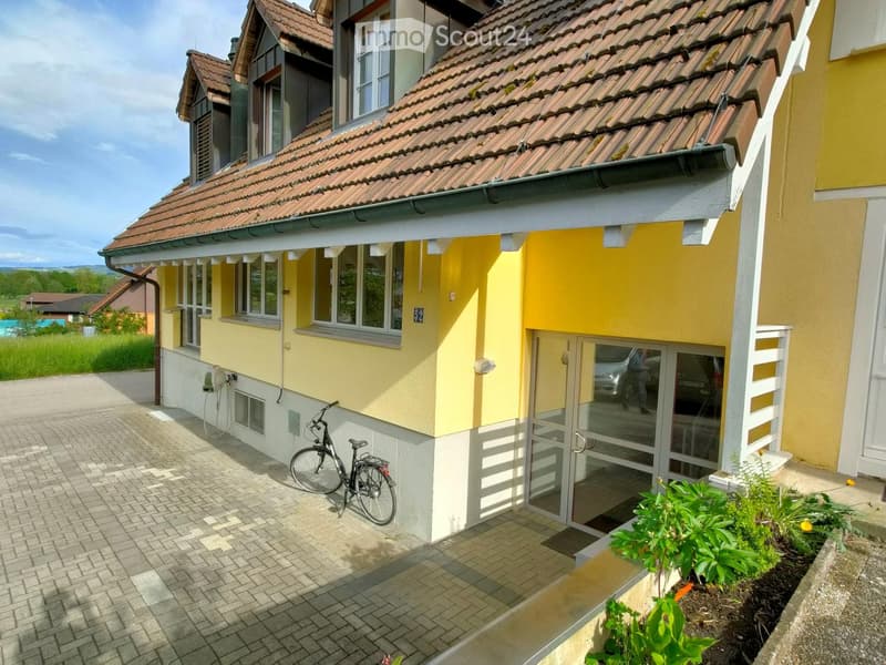 Two-story yellow house with red-tiled roof, multiple windows, and a covered entrance area. A bicycle is parked near the entrance, and there is a small garden with plants visible.