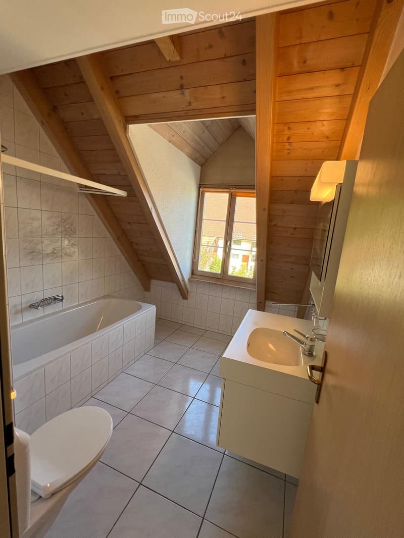 Cozy attic bathroom with wooden slanted ceilings, tiled walls and floor, bathtub, toilet, sink, and a window offering a view of the outside.