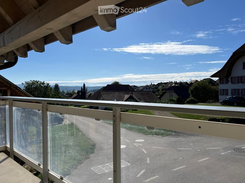 Balcony with a wooden railing and glass panels, overlooking a residential street with houses, trees, and a clear blue sky.